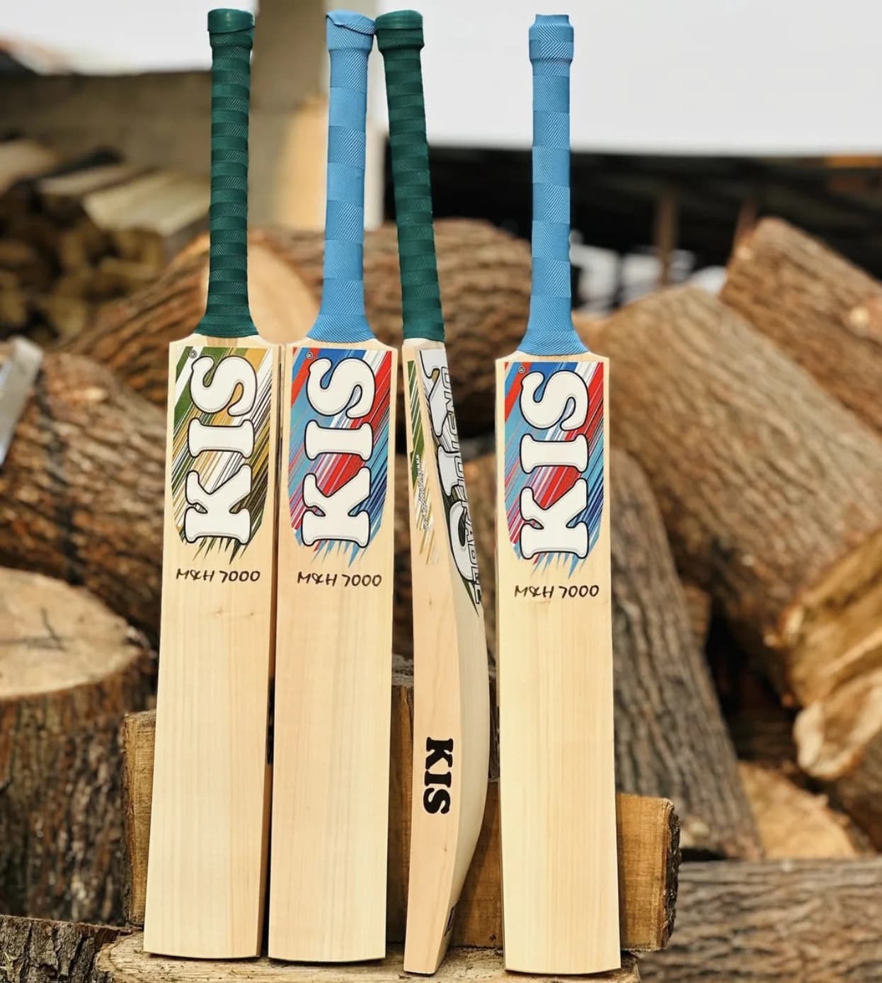 Four KIS M&H 7000 bats with green and blue grips lined up against a stack of willow logs